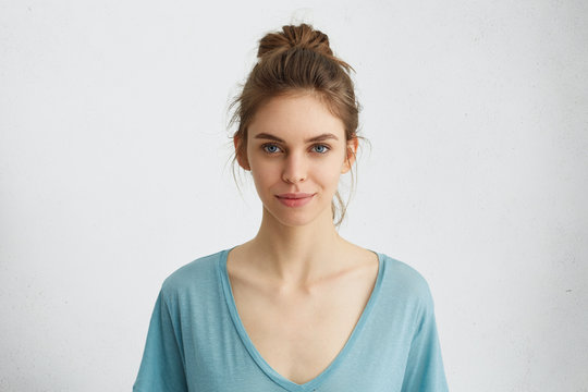 Portrait of beautiful female having blue shining eyes, thin lips, pure skin and dark hair tied in knot wearing blue loose blouse looking directly into camera having mysterious and glad expression