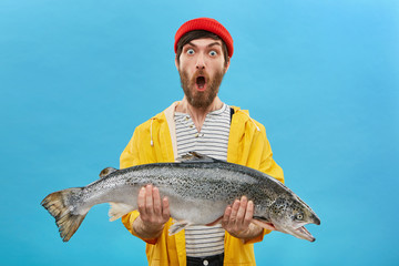 Studio shot of stylish young bearded fisherman in yellow raincoat and red hat looking in shock with jaw dropped, holding big sea-water fresh-caught fish in both hands, surprised with fine catch
