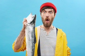 Headshot of emotional guy wearing red hat and yellow raincoat holding huge long sea fish in his...