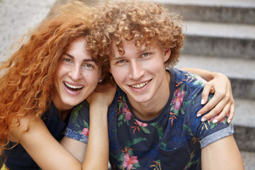 Fototapeta premium Portrait of smiling lovely couple having fun together sitting at stairs embracing each other. Happy red-haired beautiful woman embracing her boyfriend with head of curly hair. Romantic people