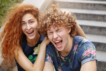Fototapeta premium Funny freckled female with reddish bushy hair scratcing head of her friend who is closing eyes and opening mouth. Couple in love making fools while sitting at stairs outdoors, laughing loudly