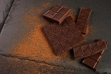 Close-up of broken chocolate pieces and cocoa powder on dark stone background with copy space.