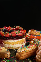 Fresh delicious colourful eclairs and summer berry cake with berries strawberries blackberries currants on dark stone background with copy space