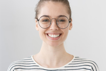 Indoor portrait of young good-looking girl in round glasses isolated on gray background wearing brown hair in bun, laughing openly at joke, expressing happiness, joy and satisfaction with life.