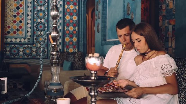 Young couple sitting on a couch in shisha cafe, arabic style, smoking and reading menu, blue walls with ornament arabic style. Shot with sony a7s on a slider. Shot with sony a7s on a tripod