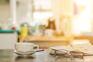 Closeup cup of hot cappuccino coffee with newspaper and spectacles on the wood table with blur kitchen room background-warm tone.