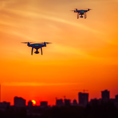 Two drone quad copters with high resolution digital camera fly aerial over spectacular sunset sky. Cityscape silhouette in the background. Modern technologies. Orange toning filter. Kiev, Ukraine