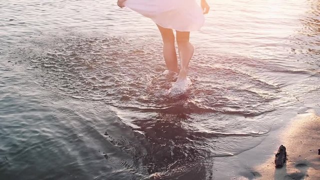 Legs of beauty girl walking barefoot along wet sand beach over sunset. Bare feet of young woman walking on beach beside ocean. Slowmotion video footage. Full HD 1920x1080p.