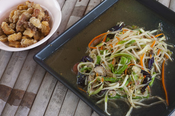Papaya salad with crab in a black tray placed on a bamboo table. and with pork snack.
