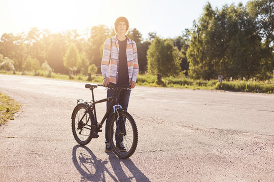 Fashionable hipster teenager wearing shirt and jeans standing on road with bicycle enjoying his hobby. Young cyclist having rest after riding bicycle. People, lifestyle and recreation concept