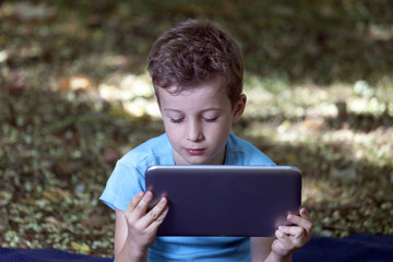 Little cute boy sitting in the park and play his favorite game on a tablet