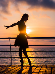 Silhouette of dancing ballerina in black ballet tutu and pointe on embankment above ocean or sea at sunrise or sunset. Young attractive blonde woman with long hair practicing stretching and exercises.