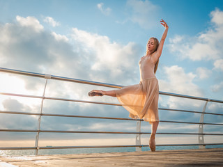 Dancing ballerina in beige silk dress and pointe on embankment above ocean or sea beach at sunrise. Young beautiful blonde woman with long hair practicing classic exercises with emotions.