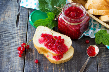 Homemade jam. A glass jar with red currant jam and white bread on a kitchen wooden table. Preserved berry.