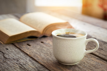 Coffee cup and open book on old wooden table with sunlight from window curtain bavkground