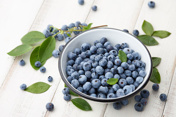 Fresh ripe blueberries with leaves in bowl on white wooden planks