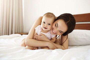Fototapeta na wymiar Happy beautiful mother in sleepwear lying on bed with her baby daughter embracing smiling.