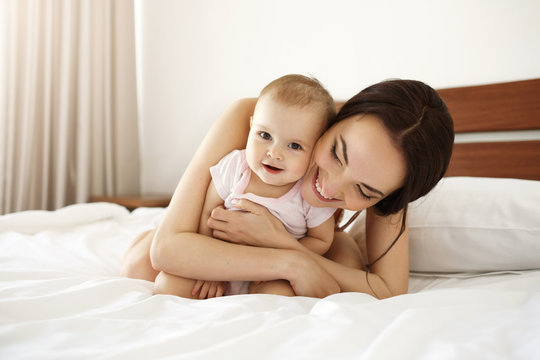 Happy beautiful mother in sleepwear lying on bed with her baby daughter embracing smiling.