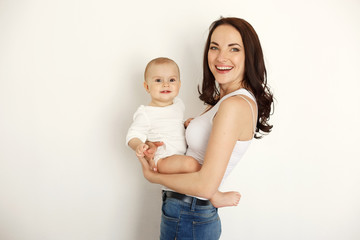 Young beautiful happy mother smiling laughing holding her baby daughter over white wall.