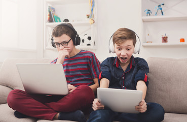 Two teenage boys with gadgets and headphones on couch at home