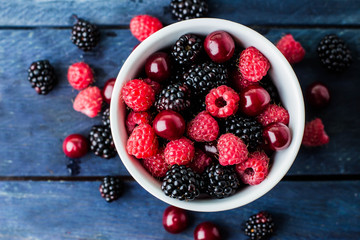Top view of white cup with Summer fresh berries on blue wooden table background. Copy space. Vegetarian food - raspberry, blackberry and cherry. Rustic style