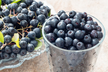 Closeup of fresh ripe black chokeberry (Aronia melanocarpa) with leaves in glass pot on brown ceramic background