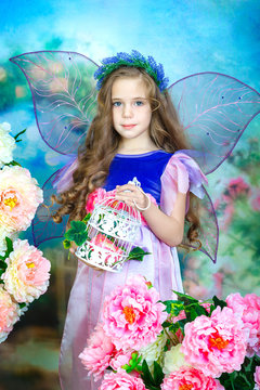 Portrait of a charming little girl with curly hair in a colorful fairy dress with transparent wings. The girl holds a decorative lace birdcage in her hands. Studio photography.