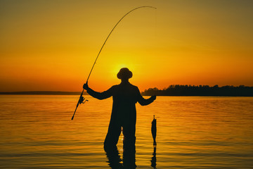 A fisherman with a fishing rod in his hand and a fish caught stands in the water against a beautiful sunset.