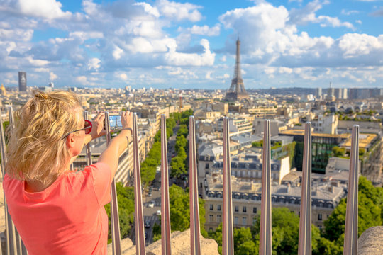 Paris skyline aerial view. Caucasian lifestyle traveler taking picture with smartphone from top of Arc de Triomphe. Blonde tourist woman takes shot of Tour Eiffel. Landmark in Paris, France, Europe.
