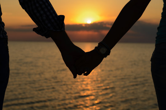 A loving couple (silhouettes of a man and a girl) hold hands in the rays of light at sunset on the sea on a pier.