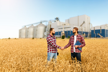 People shaking hands in a wheat field, farmer's agreement. Grain elevator terminal on background....