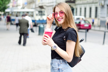 Outdoor portrait of young attractive woman with long blonde straight hair, pink glasses,  and berry's smoothie cocktail, street urban background. Healthy food detox concept. Windy hair flip