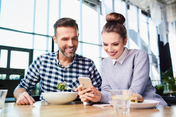 Happy middle-aged couple eating lunch together and looking at phone