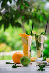 Detox water with fruits - orange and green apple and lemon and basil leaves and mint leaves on white table outdoors in summer garden. Homemade mojito or lemoade with ice cubes.