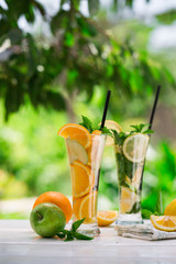 Detox water with fruits - orange and green apple and lemon and basil leaves and mint leaves on white table outdoors in summer garden. Homemade mojito or lemoade with ice cubes.