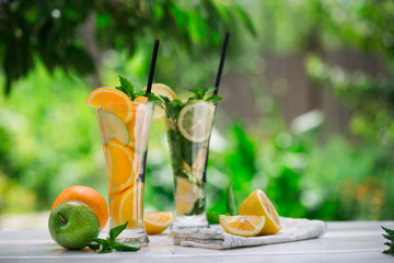 Detox water with fruits - orange and green apple and lemon and basil leaves and mint leaves on white table outdoors in summer garden. Homemade mojito or lemoade with ice cubes.