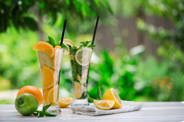 Detox water with fruits - orange and green apple and lemon and basil leaves and mint leaves on white table outdoors in summer garden. Homemade mojito or lemoade with ice cubes.