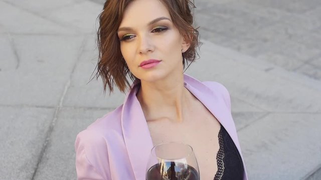 Woman with beautiful smile is standing in doorway of balcony with a glass of wine. Attractive blonde on terrace with glass of wine. Romantic girl on veranda with a wine and looking into the distance.