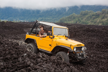 A curly-haired smiling man is sitting in the offroad yelow vehicle at the top of a valley with volcanic rock and mountains in Bali, Indonesia