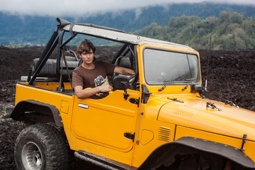 A curly-haired tattooed man is sitting in the offroad yelow vehicle at the top of a valley with volcanic rock and mountains in Bali, Indonesia © Dana Keli