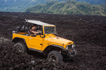 A curly-haired man is using a phone in the offroad yelow vehicle parked at the top of a valley with volcanic rock and mountains in Bali, Indonesia © Dana Keli