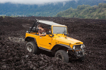 A curly-haired man in the offroad yelow vehicle is looking backward at the top of a valley with volcanic rock and mountains in Bali, Indonesia © Dana Keli