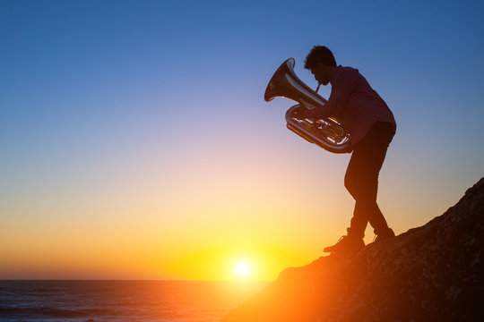 Tuba - instrument. Silhouette of a young man playing the trumpet on rocky sea coast during sunset.