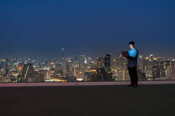 Fototapeta na wymiar Asian technician engineer standing and using the laptop over the cityscape background at night time, Business success and technology concept