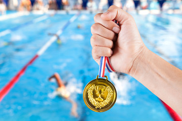 handful of asian man holding gold medal with blurry background of swimming pool.