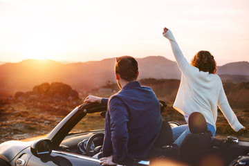 A lovely couple have fun in a convertible at sunset, around them beautiful nature - obrazy, fototapety, plakaty