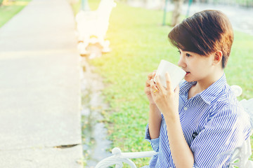 Beautiful short hair woman sitting on a white chair and drinking a favorite beverage in white cup for resting between her outdoor working business in the park.
