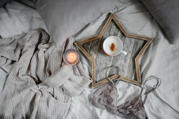 A cup of coffee and a candle in a Scandinavian wooden tray in a cozy bed with pillows.