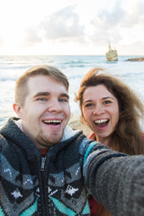 Young laughing couple hiking taking selfie with smart phone. Happy young man and woman taking self portrait with sea or ocean scenery on background. Winter time