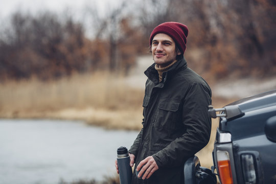 Smiling man holding insulated drink container while standing by sports utility vehicle at lakeshore